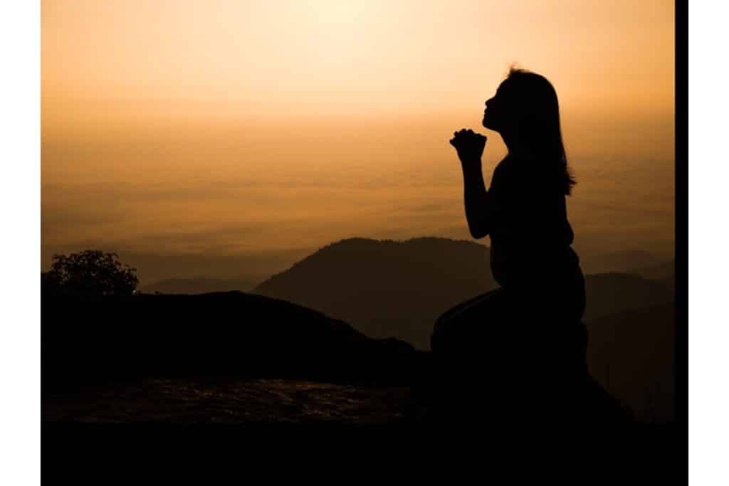 devant un coucher de soleil on devine une silhouette d'une femme qui prie agenouillée devant un paysage de montagnes, avec les mains jointes.