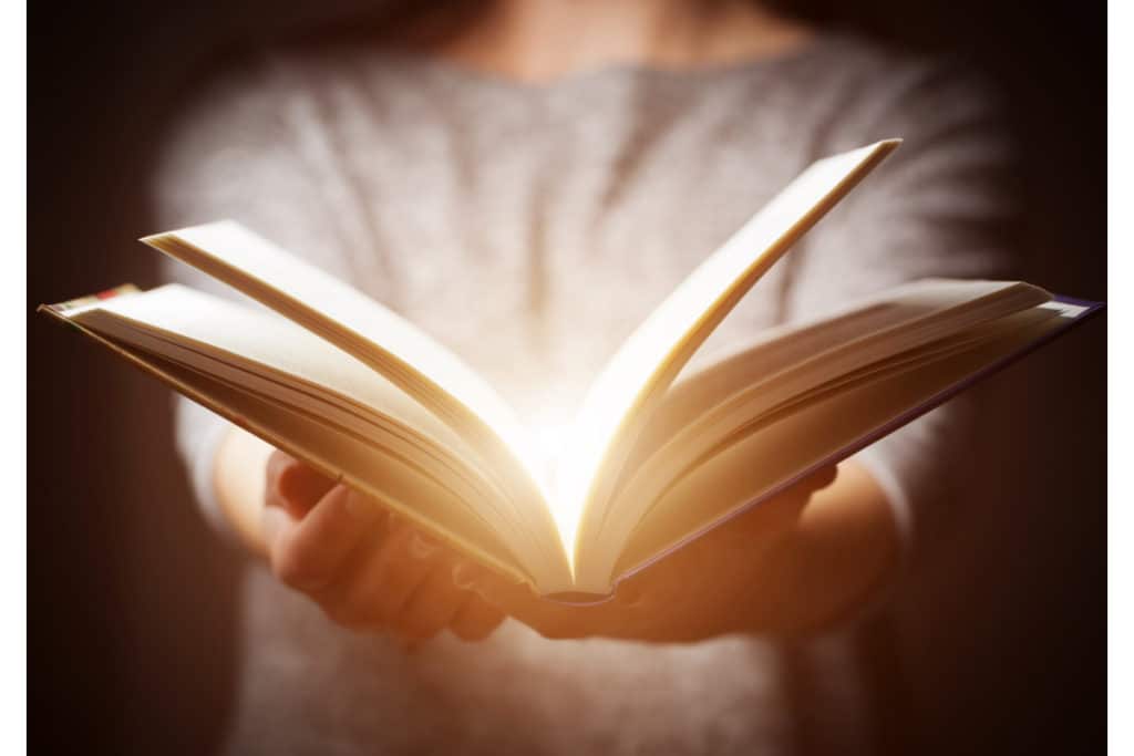 une personne tient un livre ouvert entre ses mains. Une lumière jaillit de milieu du livre.
