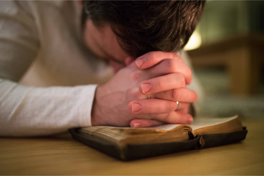 un homme a les mains croisées en prière sur un vieux livre. sa tête est posée sur ses mains.