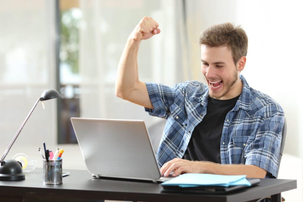 un jeune homme est assis devant un ordinateur portable à un bureau et lève le poing droit vers le haut en souriant très largement la bouche grande ouverte.