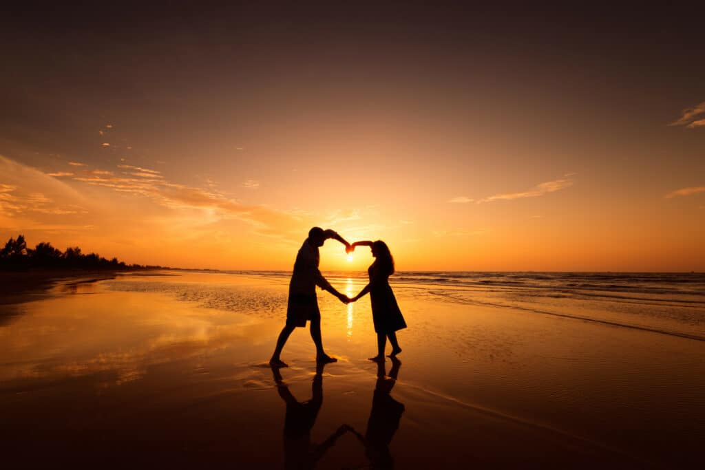 un homme et une femme devant un coucher de soleil sur une plage avec les bras qui forment un coeur. prière pour attirer l'amour d'un homme.