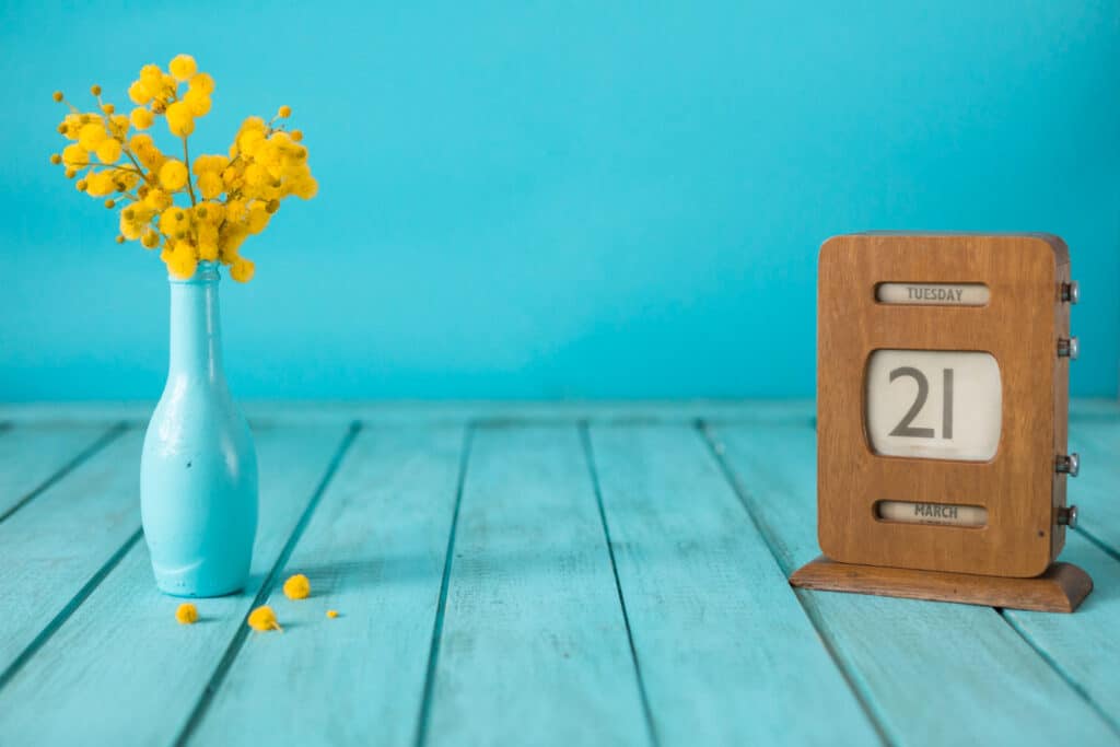 un calendrier en bois à côté d'un bouquet de mimosa jaune dans un vase bleu ciel. Les deux sont posés sur un parquet avec un fond bleu ciel