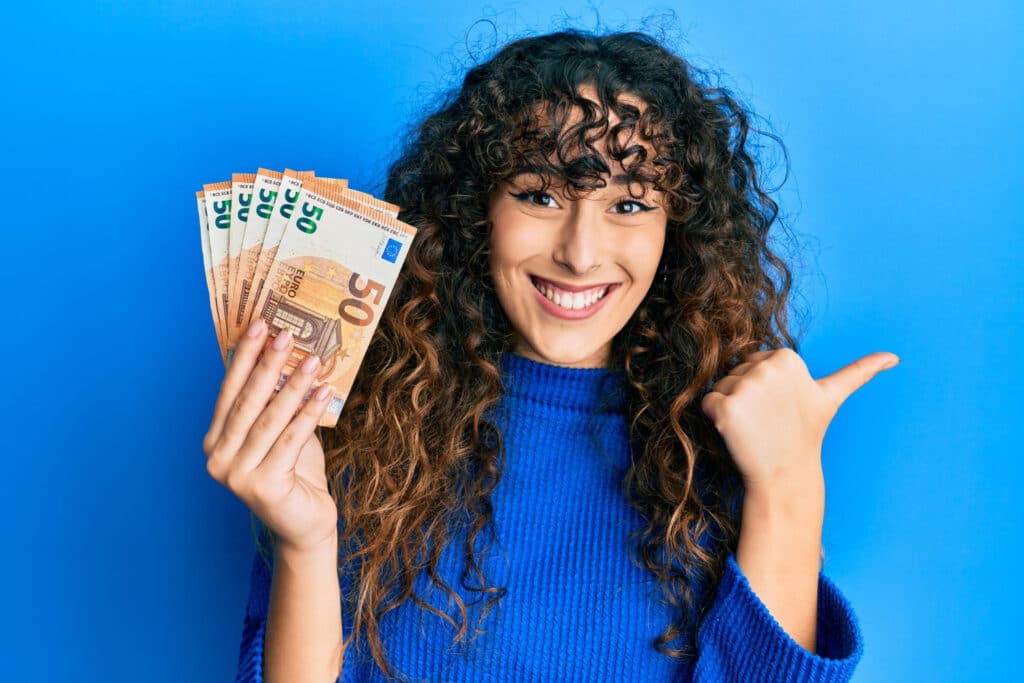 jeune femme brune aux cheveux bruns en pull bleu qui tient des billets de 50 euro et qui sourie; devant un fond bleu.