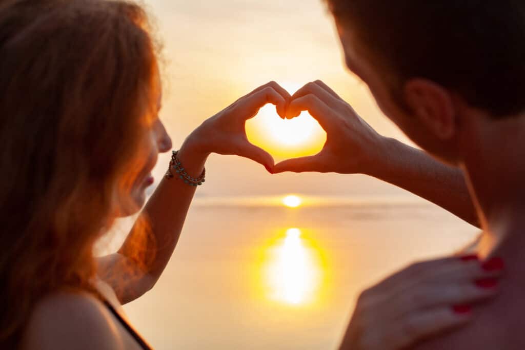 un couple d'homme et femme qui forment un cœur avec leurs mains devant un coucher de soleil