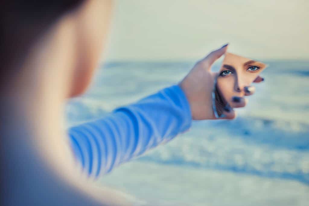 vue de dos floutée d'une femme en haut à manches longues bleu qui tient un bris de miroir et qui regarde son reflet, avec un arrière plan bleu clair.