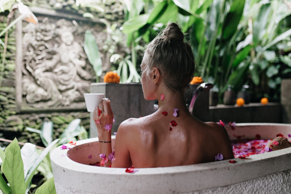 une femme vue de derrière dans un bain avec des pétales de fleurs roses, avec des plantes vertes et un mur en pierre en arrière plan